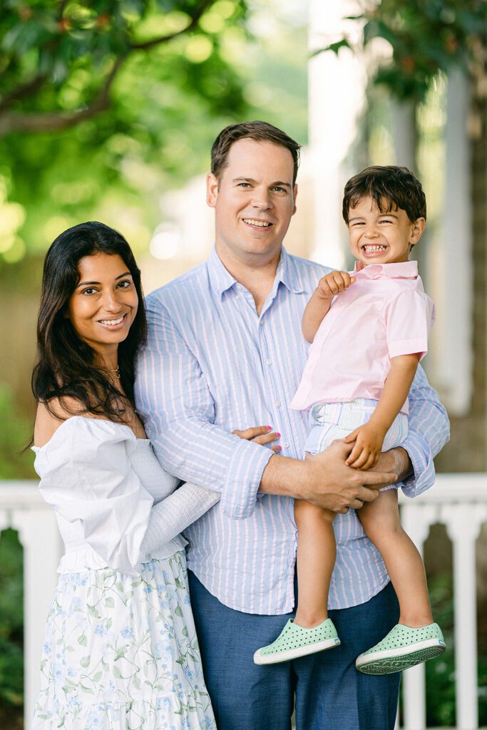 Mom, dad, and brunette toddler boy are smiling at the camera, all dressed in shades of blue and green, with a pop of pink from the toddler's shirt. 