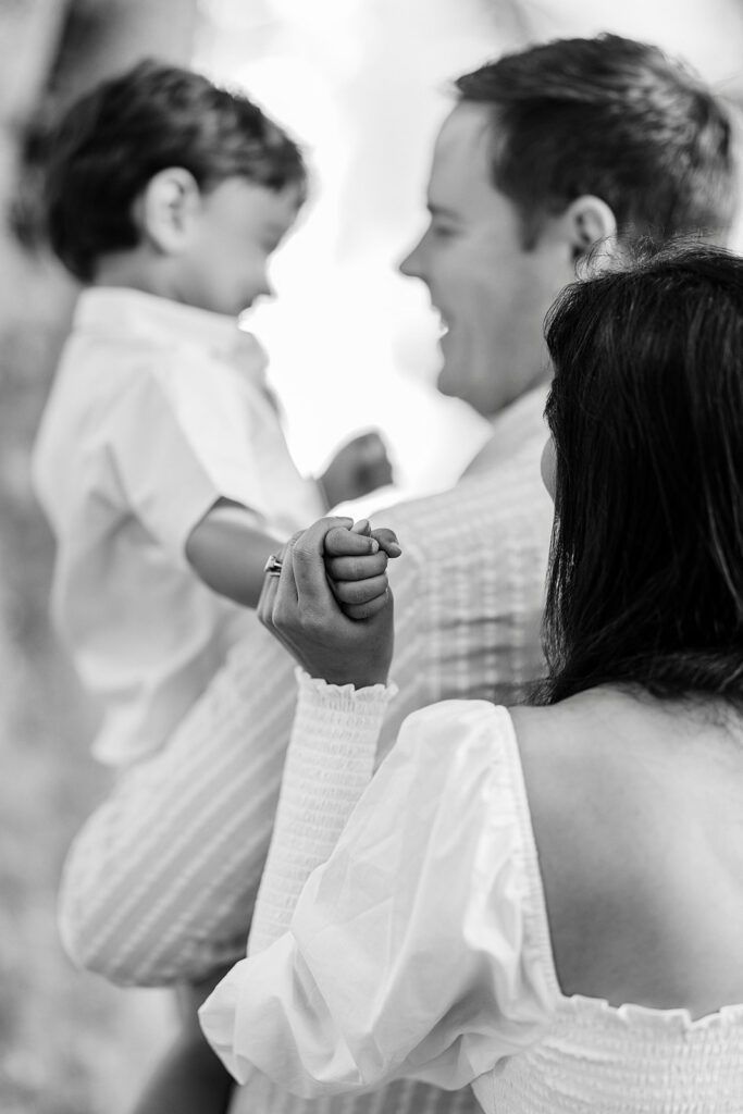 Dad is holding toddler boy with mom standing behind them holding onto toddler boy's hand. Dad and toddler boy are smiling at each other. 