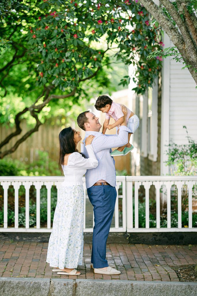 Brunette toddler boy is being hoisted up in the air by dad who is smiling up at him, while mom stands behind dad with her hand on his shoulder smiling up at the toddler boy as well. They are standing on a brick sidewalk with a white bench behind them. 