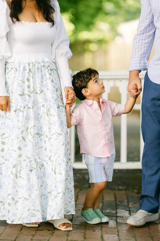 Brunette toddler boy in pink button down shirt and pastel striped shorts is standing between mom and dad holding both of their hands, while looking up at dad. 