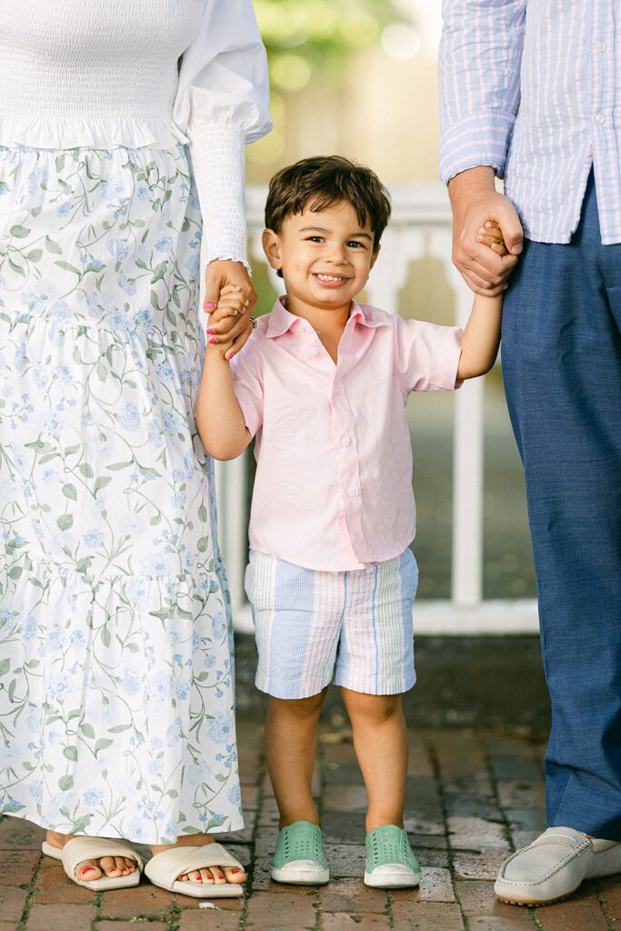 Brunette toddler boy in pink button down shirt and pastel striped shorts is walking in between mom and dad holding both of their hands with a big smile on his face.