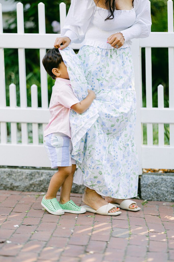 Mom and brunette boy are standing on a brick walkway in front of a white fence, with mom lifting her long skirt to cover the boys face as he hides and plays. 