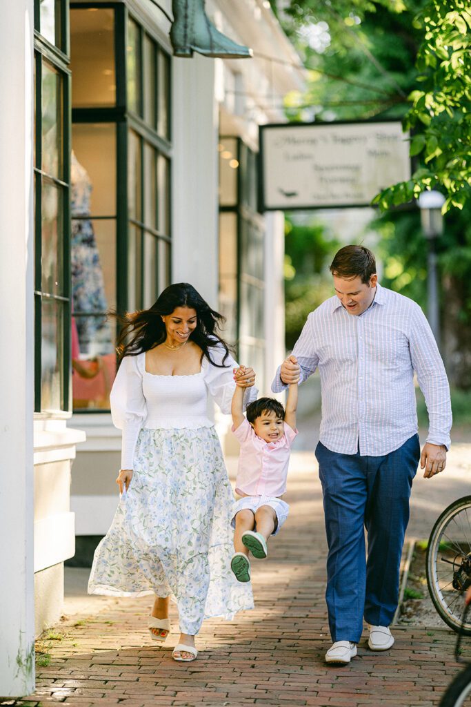 Brunette toddler boy in pink button down shirt and pastel striped shorts is holding onto both mom and dad's hands and being swung into the air while walking along a brick sidewalk.