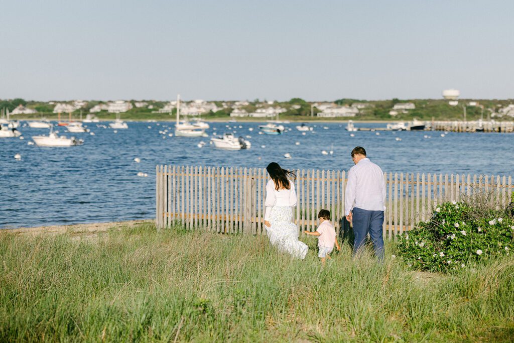 Mom, brunette toddler boy, and dad are walking in the grassy dunes towards the beach, the ocean and many sail boats visible in the distance. 