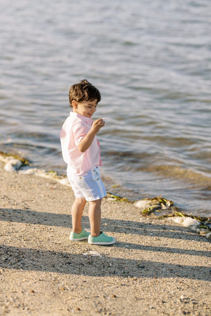 Brunette toddler boy in pale pink button down shirt is smiling at a seashell in his hand as he stands on the sand with soft waves coming in right in front of him. 