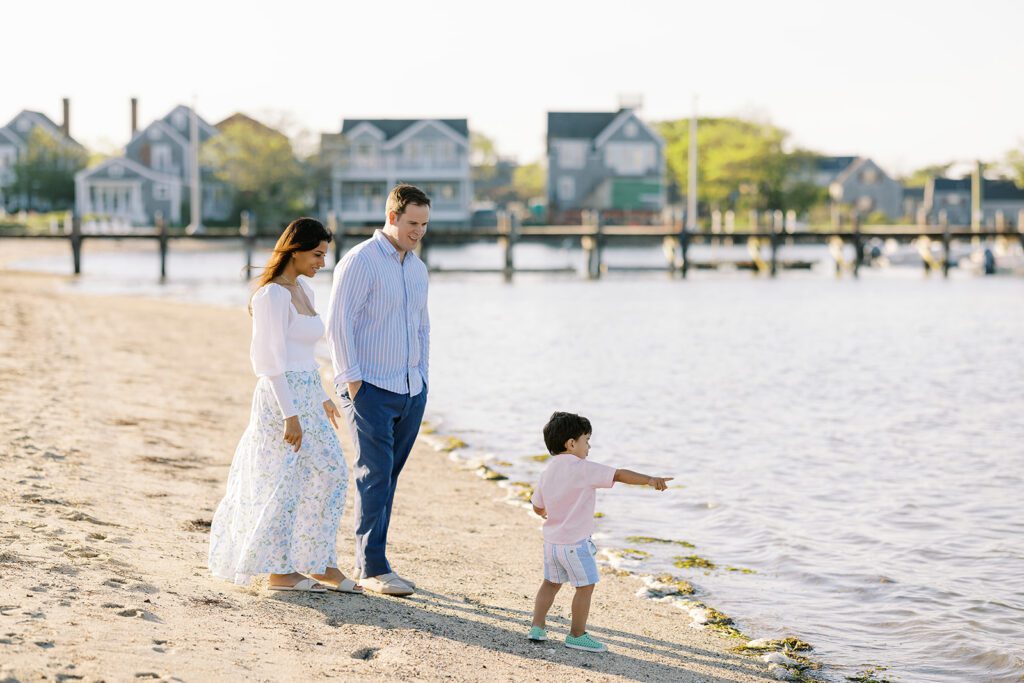 Brunette toddler boy is standing along the shoreline pointing out something in the water ahead of him. Mom and dad are smiling lovingly at him from behind.