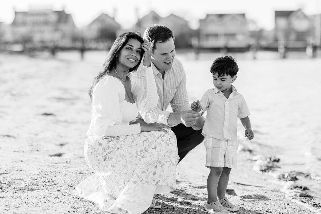 Mom and dad are crouching down on the beach beside a brunette toddler boy who is showing them a seashell. Dad is looking down at the treasure and mom is smiling while moving hair behind her ear. 