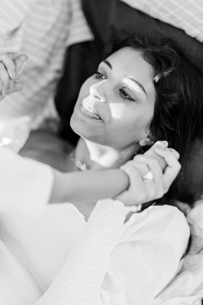 Mom is laying down on dad's leg and holding onto toddler's hands with a mouth-closed smile.