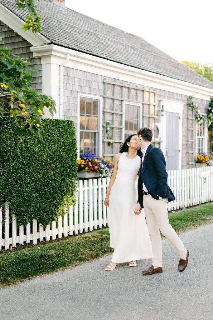 An engaged couple holds hands and steals a kiss walking alongside a grey shingled cottage adorned with flowers and greenery.