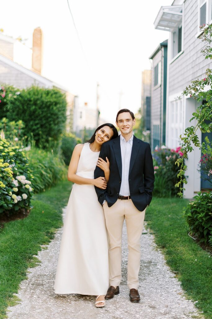 Brunette woman in an ivory dress is clutching the arm of a brunette man in a navy sport coat and khaki's, both are smiling and she is resting her head on his shoulder. 