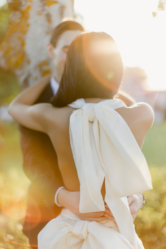 Woman in a white dress, photographed from the back to show off the large bow details on her gown, puts her hands around her partner's neck while he leans against a tree and pulls her closer by the waist. Lens flare gives the image a warm and magical vibe.