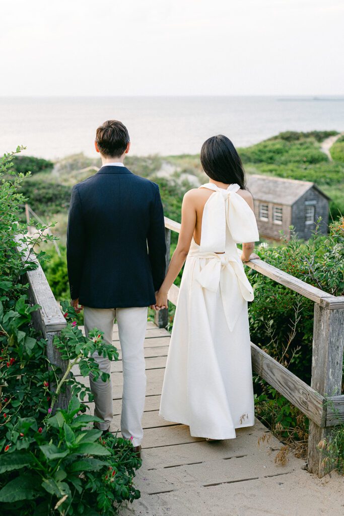 A couple stands at the top of a set of wooden stairs leading down to the beach on the island of Nantucket, taking in the view of the dunes and ocean below.