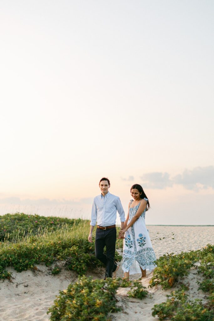 Man in a light blue button down and dark slacks is holding the hand of a brunette woman in a white, blue, and green floral block-print sundress as they walk in the sandy dunes. 