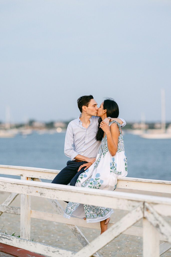 Man in a light blue button down and dark slacks is kissing a brunette woman in a white, blue, and green floral block-print sundress as they sit on top of a white wooden fence on the beach.