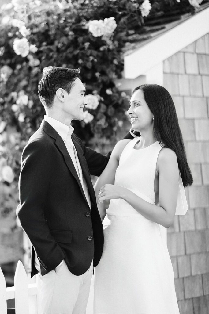 Brunette woman in ivory dress and a man in a sport coat are standing closely, facing one another, and smiling. 