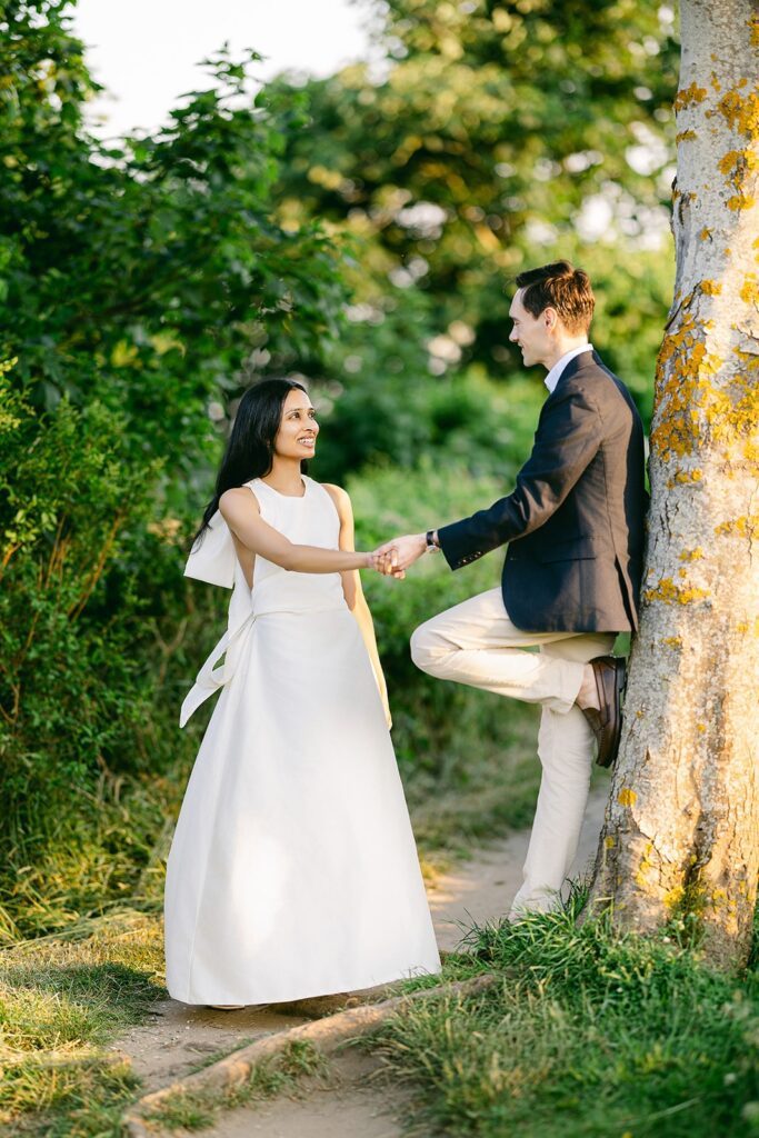 A man in a sport coat is leaning against a tree with one leg bent, holding the hand of a brunette woman in ivory dress who is smiling at him. 