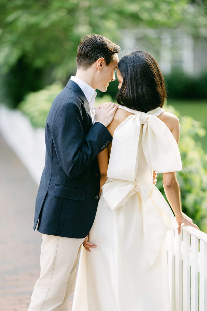 Brunette woman in ivory dress is standing in front of a white fence with a man in a navy sport jacket and khaki pants right beside her leaning towards her - he's resting his hand on her shoulder and their foreheads are almost touching. 