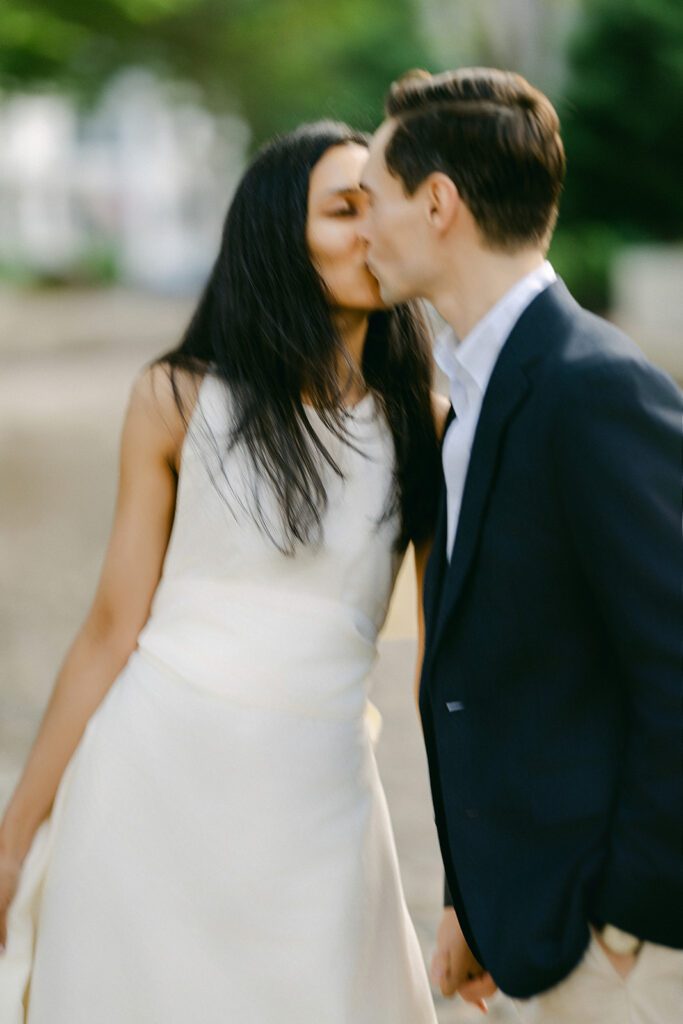 Brunette woman in ivory dress and a man in a sport coat are holding hands & kissing one another, eyes closed. 