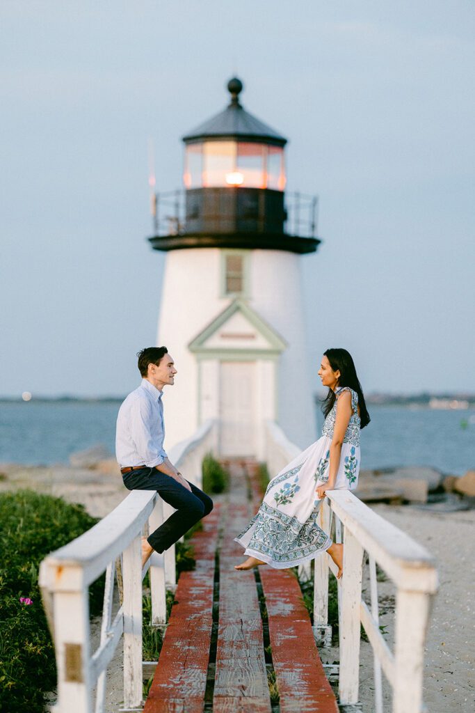 Man in a light blue button down and dark slacks & a brunette woman in a white, blue, and green floral block-print sundress each sit atop a white railing smiling at each other. The pathway between them leads to a lighthouse visible in the background. 