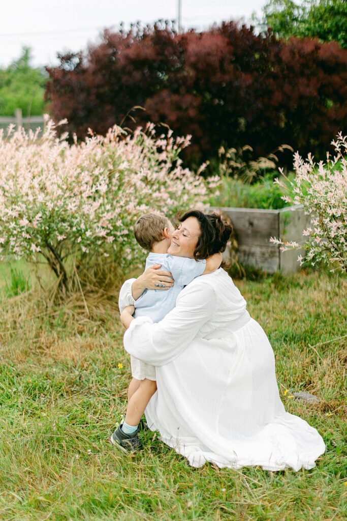 Brunette mom in a white long sleeve dress is knelling down in the green grass and giving a her brunette toddler boy a big embrace. The boys arms are wrapped around mom's neck. 