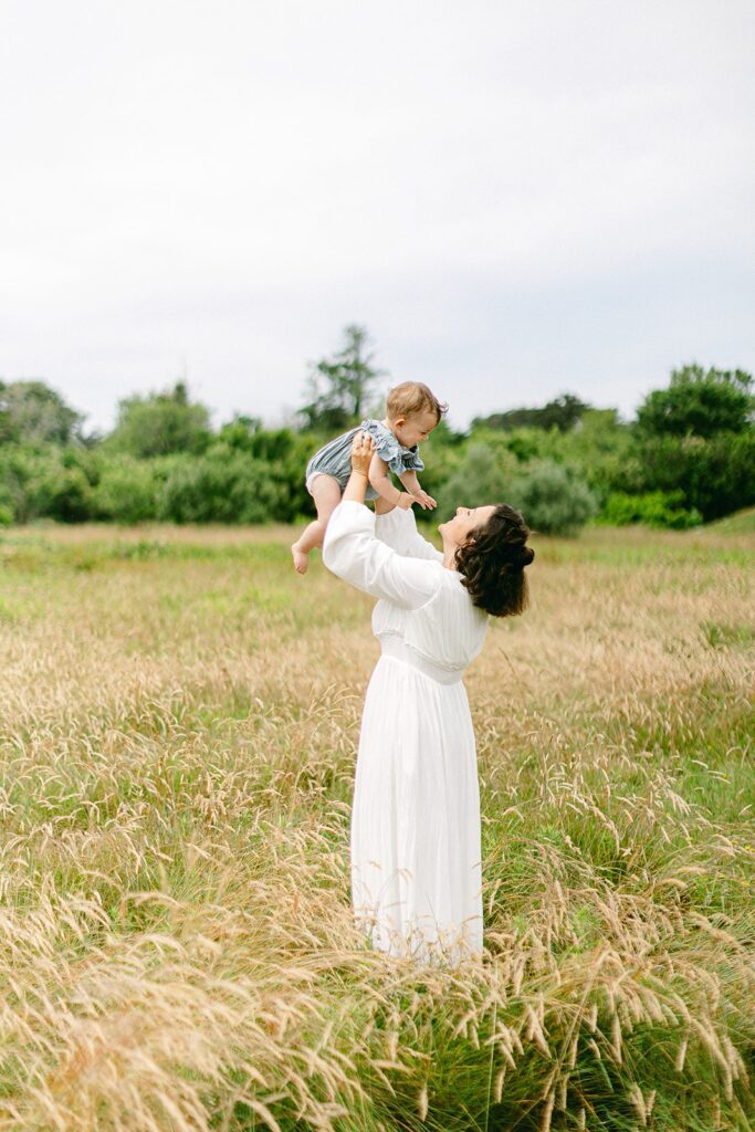 Brunette mom in a white dress is standing in a field and holding a baby up in the air, smiling at her. 