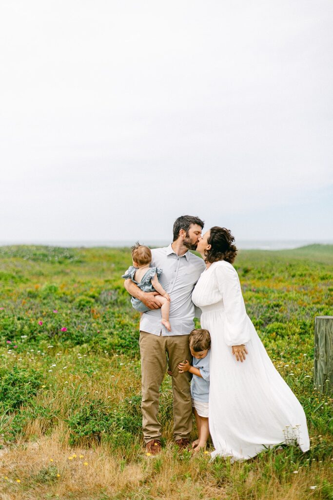 Dad and Mom are standing side-by-side in a field and kissing. Dad is holding a baby girl and their toddler boy is standing in between them. 