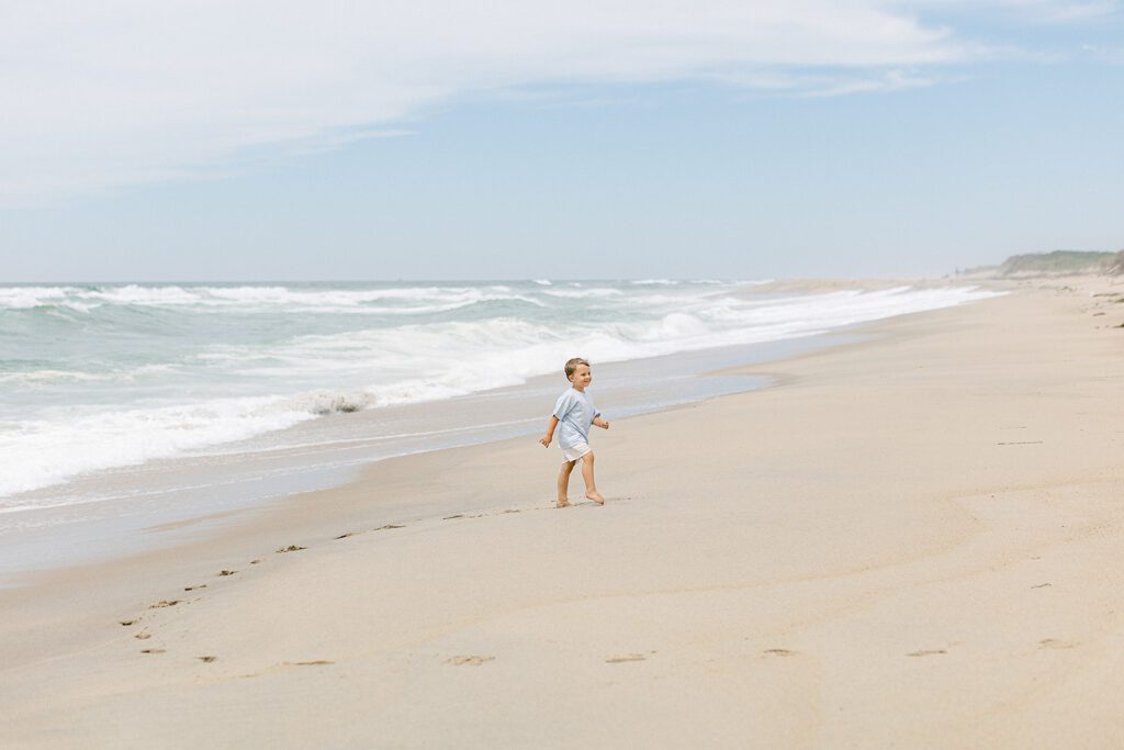 Toddler boy walking on the beach with the waves crashing and behind him along with a trail of his footprints in the sand. 