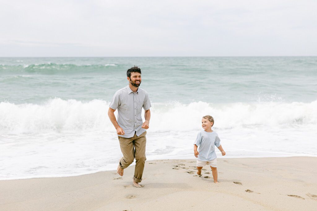 Bearded dad in a short sleeve button-down and brown pants is running with his toddler boy on the beach away from the water. Toddler boy is smiling up at dad. 