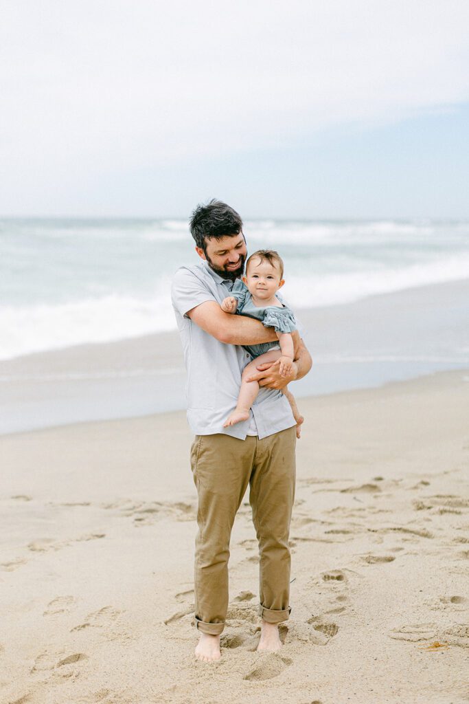 Bearded dad is standing on the beach holding his baby girl and smiling down at her. 