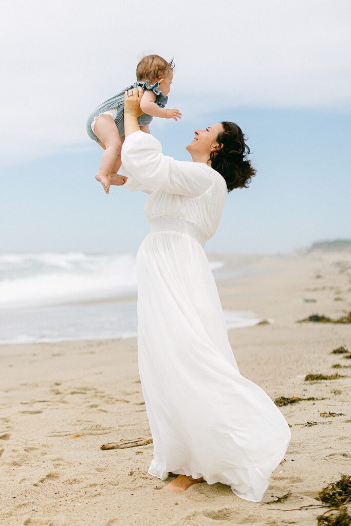 Brunette mom in a long sleeve, flowing, white dress is standing on the beach holding her baby girl up in the air and smiling. 