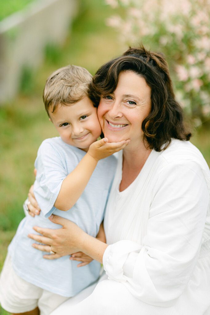 Brunette mom in a white dress is posed next to a sandy brown toddler boy - her arm I around him and he is cupping her chin with this hand. they are both smiling. 