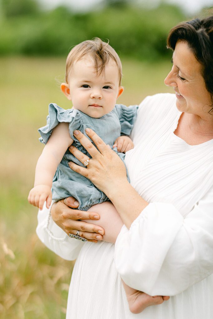 Brunette mom in a white long sleeve dress is holding a brunette baby girl on her hip who is wearing a denim romper with ruffle sleeves and staring straight ahead.  