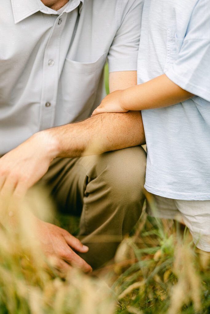 Close up view of a toddler boy resting his hand on his dad's arm. Dad is crouched down with his arm resting on his bend knee. 