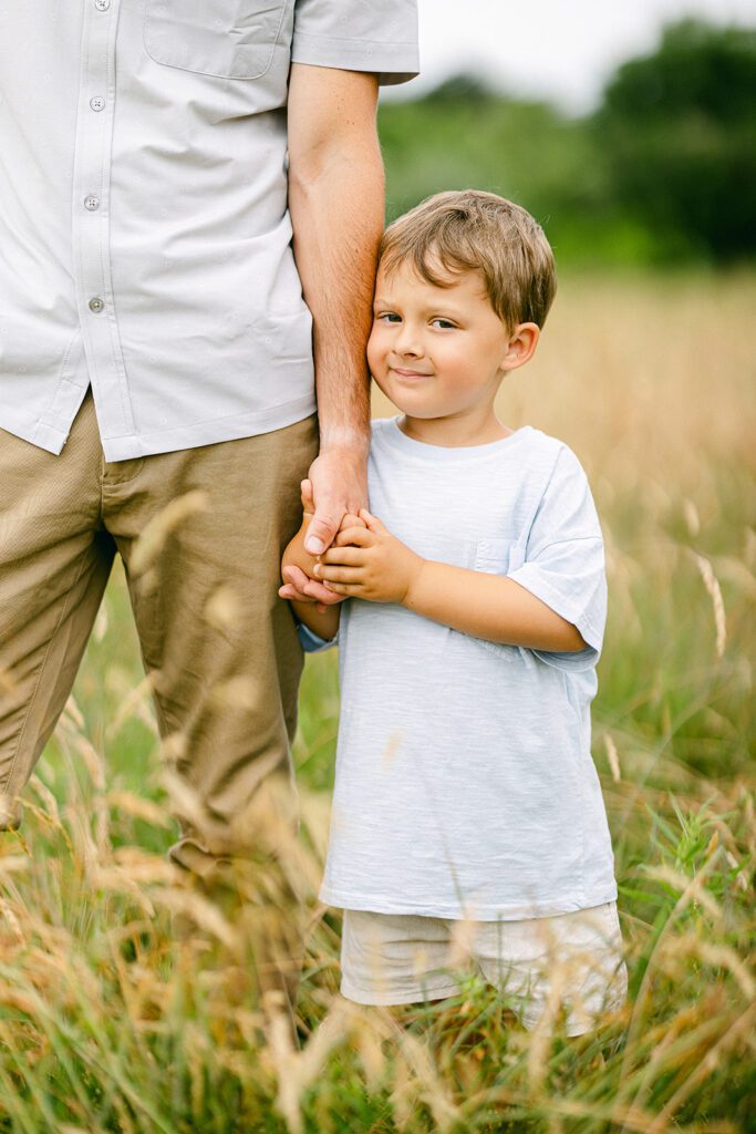 Brunetter toddler boy is grasping onto dad's hand with both of his hand, his cheek touching dad's forearm, with a little smile on his face. 