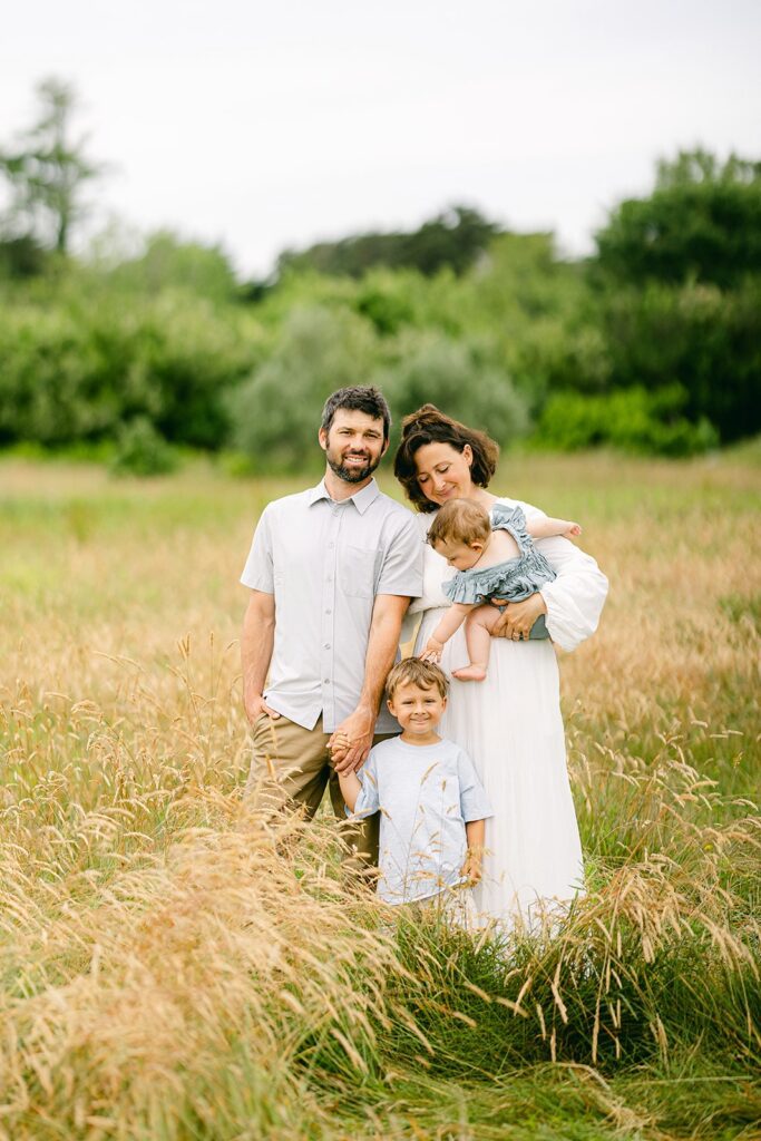 Dad and Mom are standing side-by-side in a field. Dad is holding toddler boy's hand and he is smiling. Mom is holding baby girl who is reaching down to touch the toddler boy's head. Mom is smiling down at the baby. 