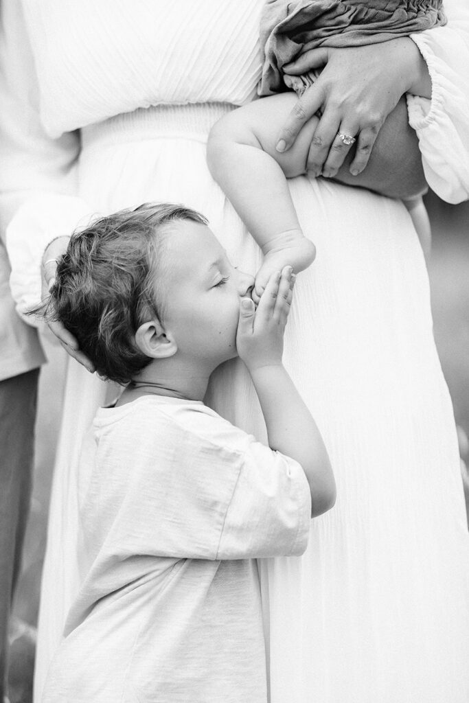 Mom in dress is holding a baby on her hip, while a toddler boy is gentry holding the babies foot and kissing it, eyes closed. 