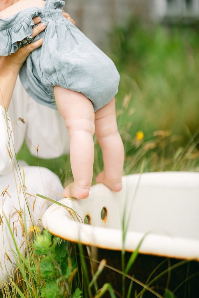 Mom is helping her baby stand up on the ledge of a bathtub that is outside in the grass. The baby is wearing a denim romper and nothing else. 