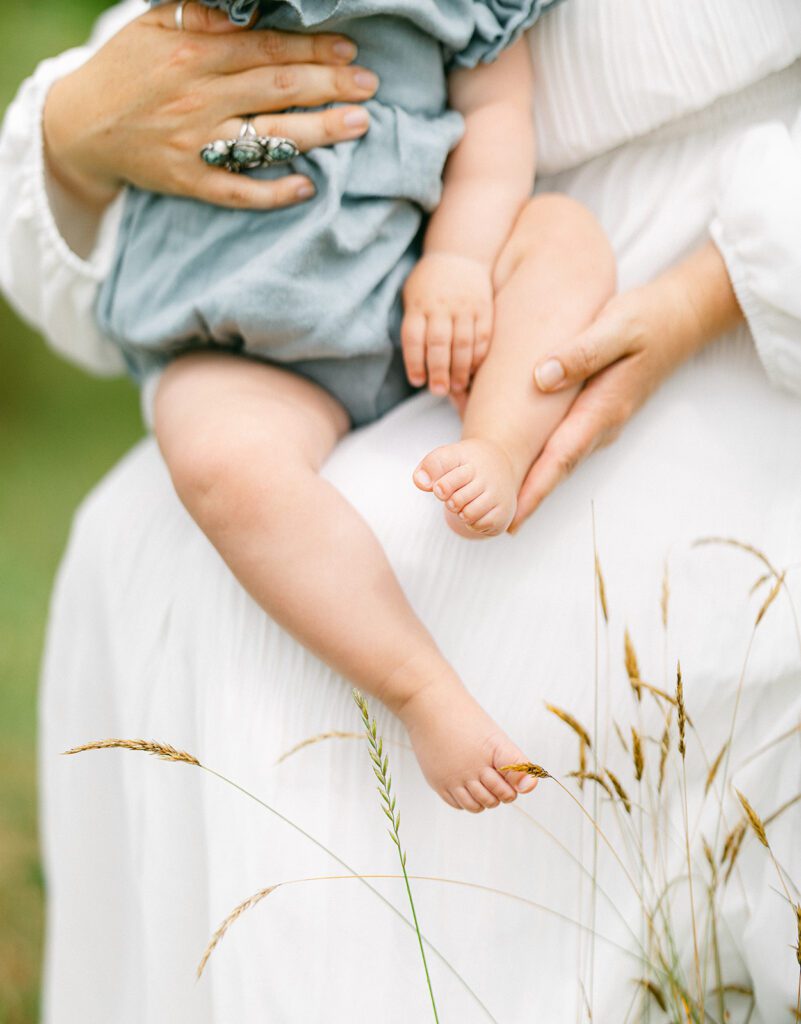 Mom in a white dress is holding a baby girl on her hip who is wearing a denim romper. Mom is gently holding her leg with one hand and a big silver and turquoise ring is visible on the other hand. 