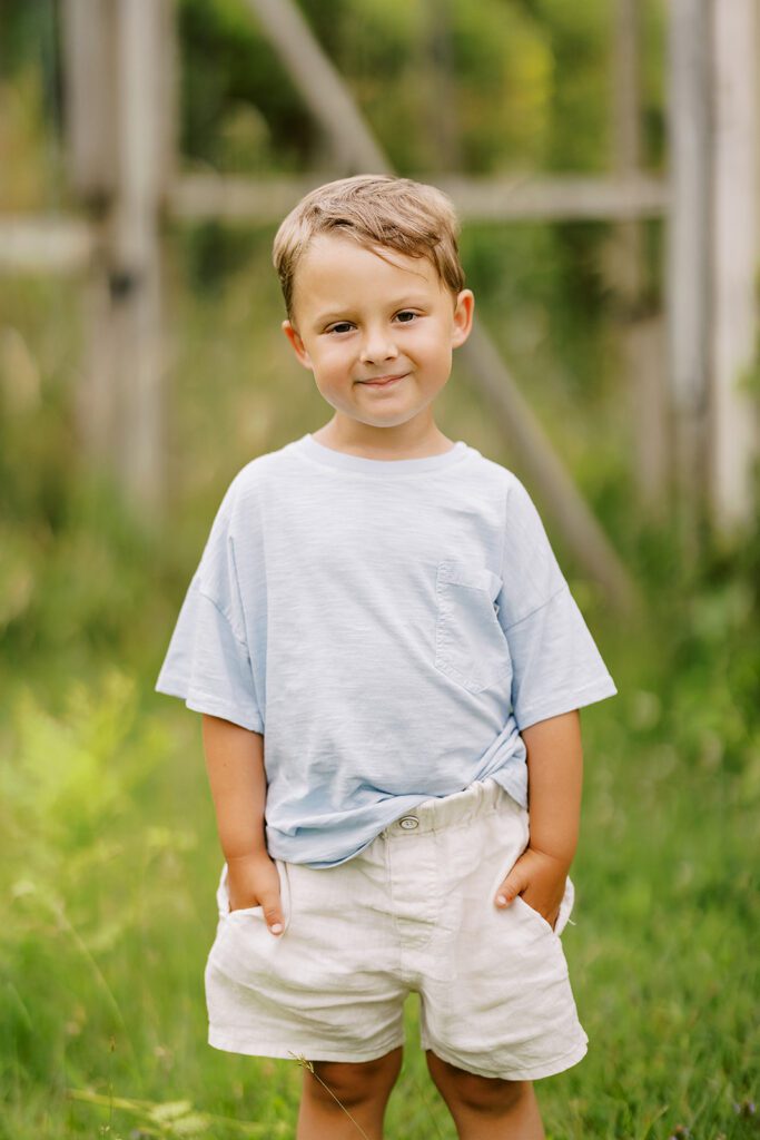 Brunette toddler boy is wearing a light blue t-shirt and ivory shorts - both of this hands are in his pockets and he has a little smile on his face. 