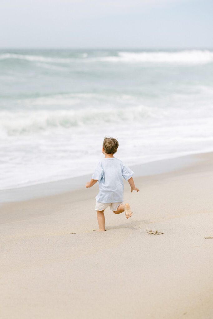 Toddler boy is running on the beach towards the ocean where the waves are crashing. 