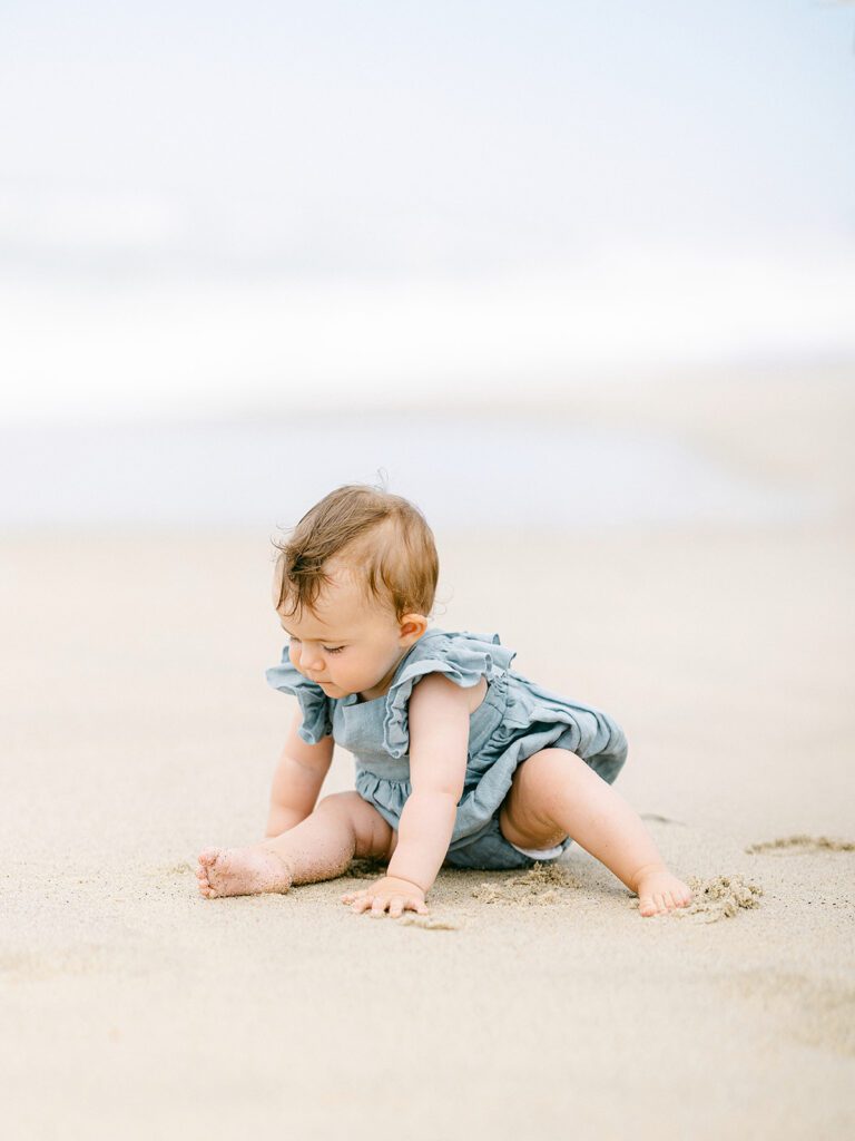 Brunette baby girl wearing a chambray romper with ruffled sleeves is sitting in the sand and exploring. 