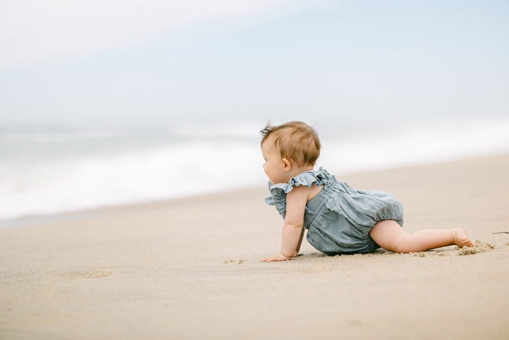 Brunette baby girl is crawling on the sand towards the ocean wearing a chambray romper with ruffles on the sleeves. 