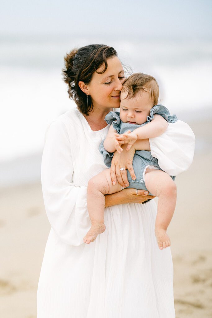 Brunette mom in a long sleeve, flowing, white dress is standing on the beach holding her baby girl close to her chest, with her cheek resting on her baby's head. 