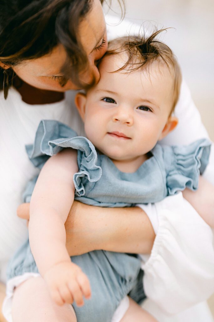 Brown-eyed baby girl is staring straight ahead with a hint of a smile on her face. Mom is seen kissing her head. 