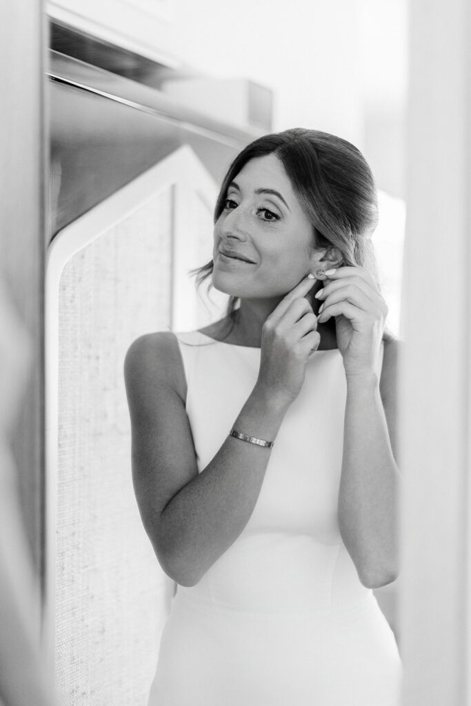 A women in a white boatneck, sleeveless dress is putting her earrings on. A Cartier love bracelet is resting on her wrist and she's smiling at herself in front of a mirror. 