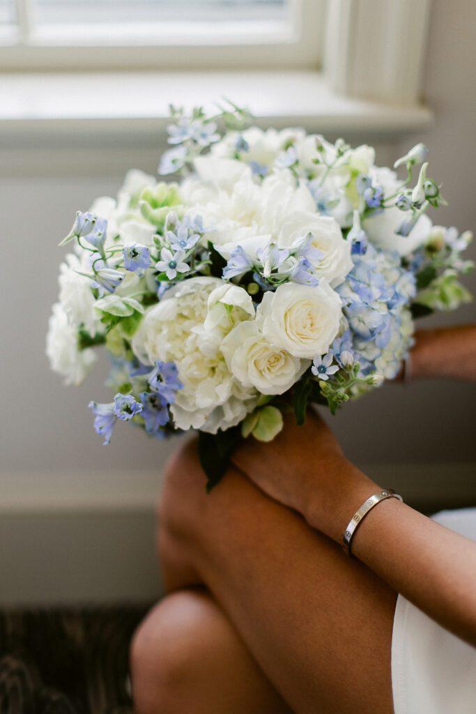 A woman is holding a bouquet of white roses accented with light blue flowers and greens. A gold Cartier love bracelet is adorning her wrist and she's resting the bouquet on her knee. 