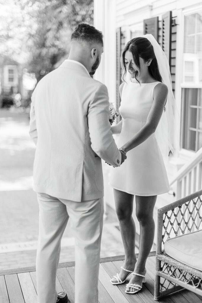 A groom wearing a suit is holding the hands of a bride in a boatneck, sleeveless mini dress on a porch. 