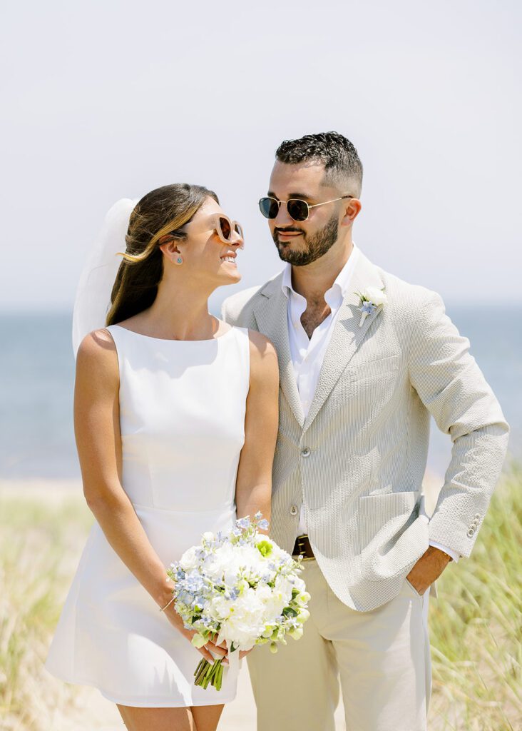 A brunette bride in a white, boatneck, sleeveless mini dress has sunglasses on and is smiling up at her brunette, bearded groom who has one hand in the pocket of his neutral colored suit and is wearing sunglasses as well. 