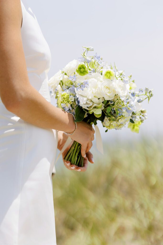 A woman in a white dress is standing with her arms at waist-height and is soldering a bouquet of white, blue, and green flowers. 