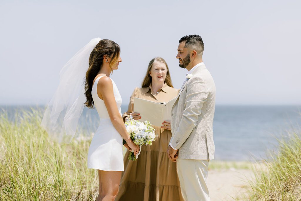 A brunette bride in a white sleeveless mini dress is standing opposite her brunette, bearded groom wearing a neutral suit. The efficient is standing between them in a brown dress holding open a binder. 